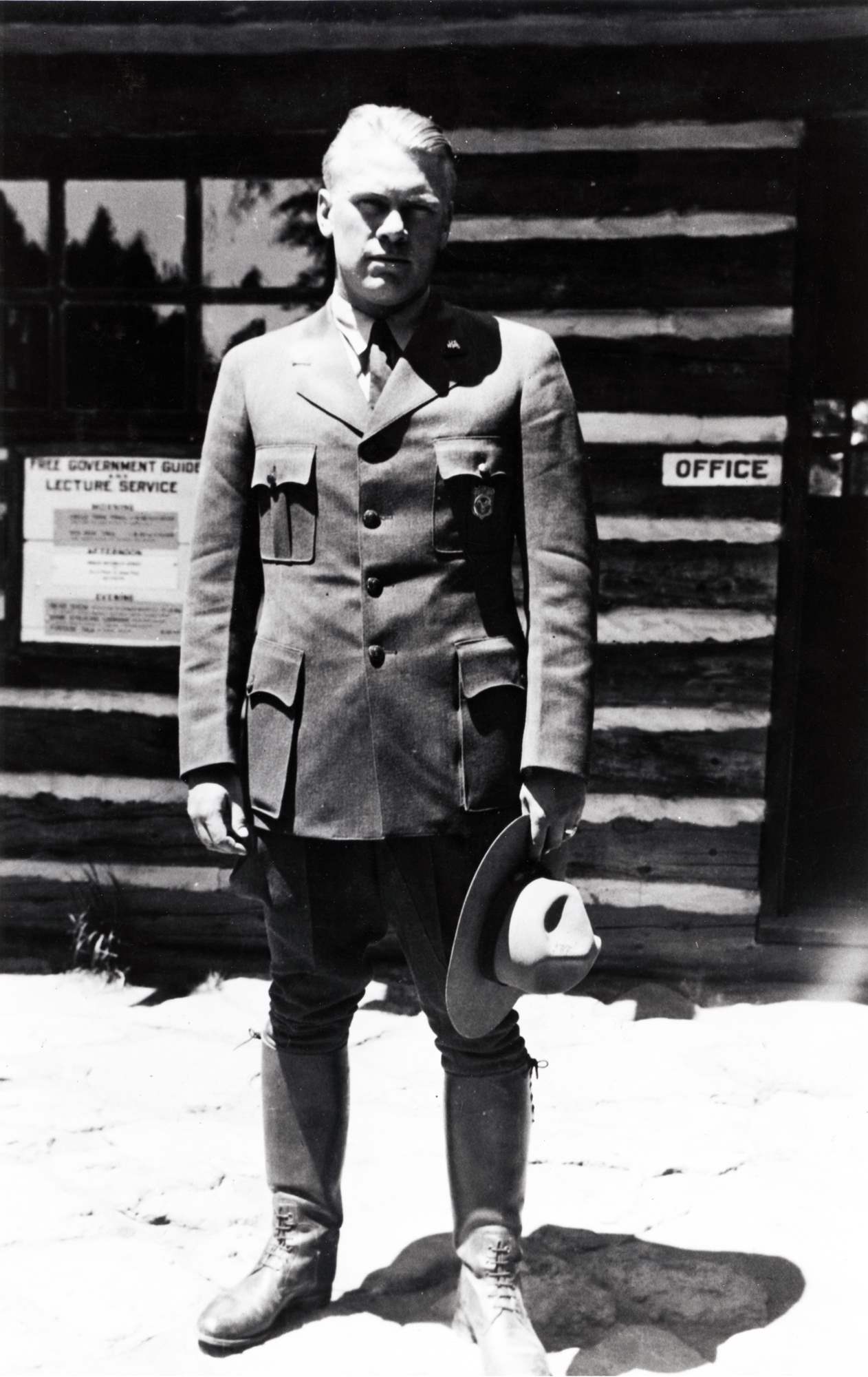 Gerald Ford poses in front of a log building while wearing the NPS uniform with shield-shaped badge and holding a broad brim hat.